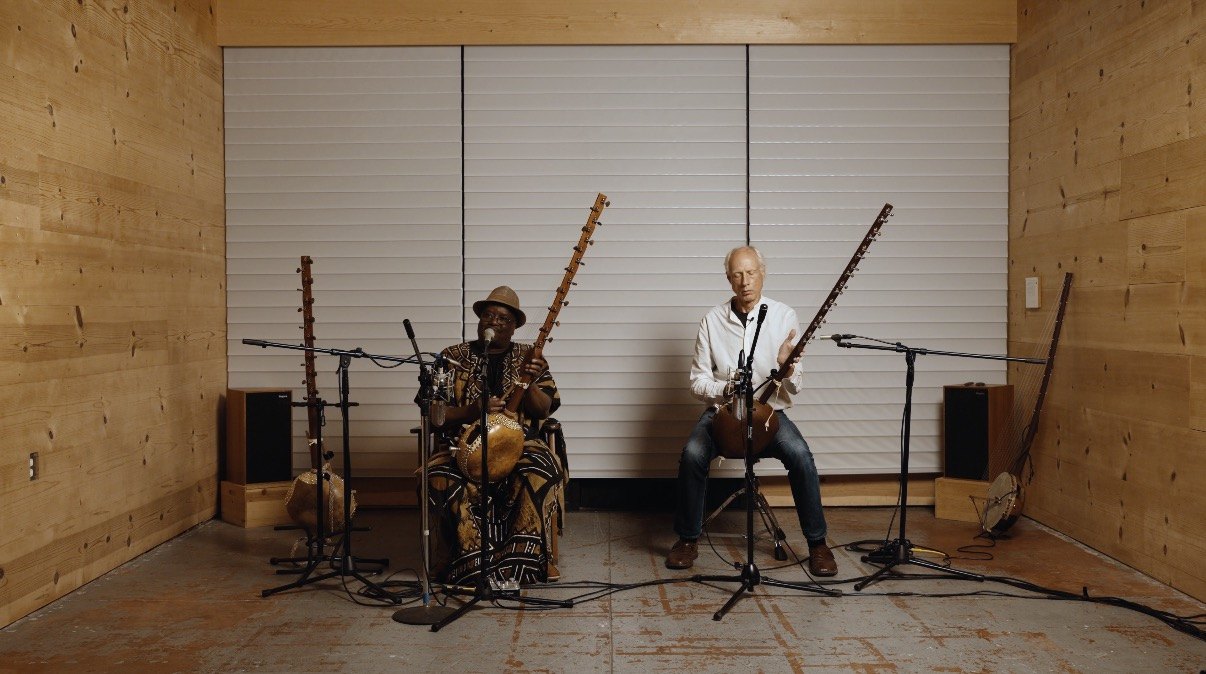 Two musicians playing koras side by side in a wood-lined room