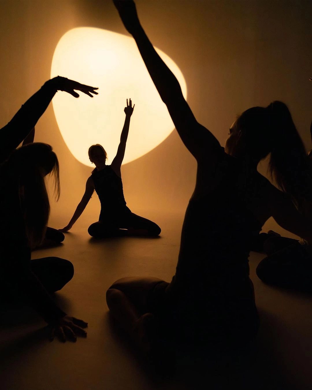 Dancers in silhouette reaching toward a soft disc of warm light
